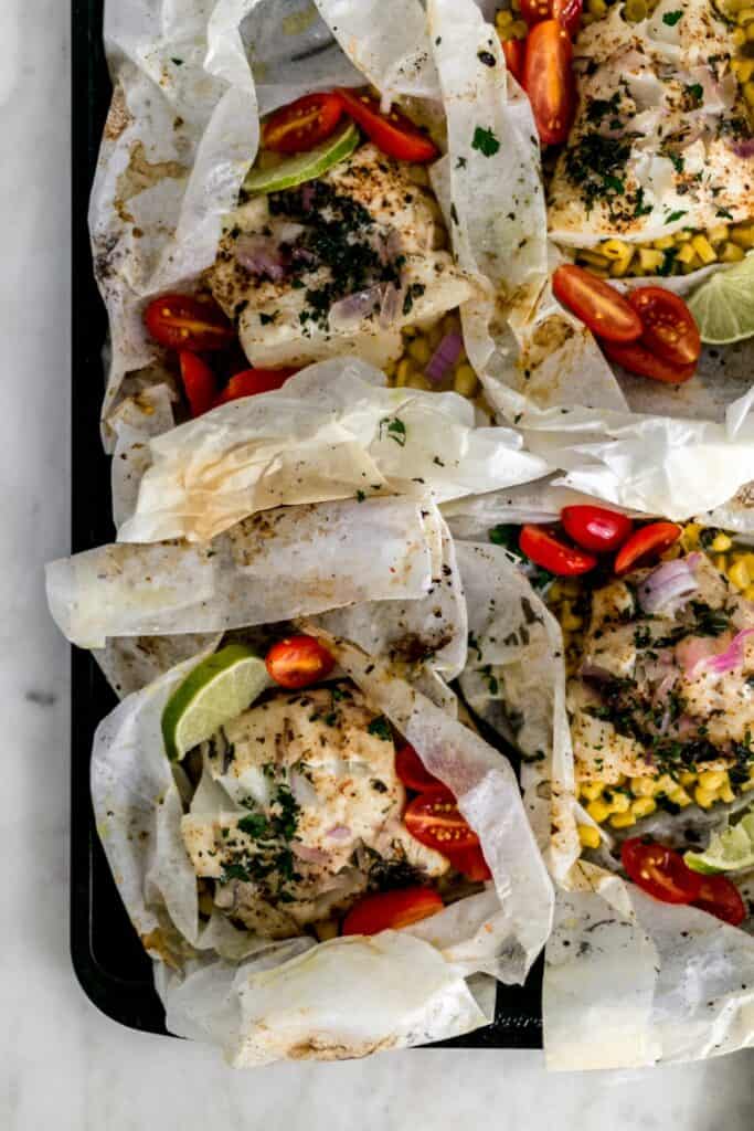 baked cod packets in parchment paper on baking sheet. Packets have fish, corn, red onion, tomatoes, and lime slices.