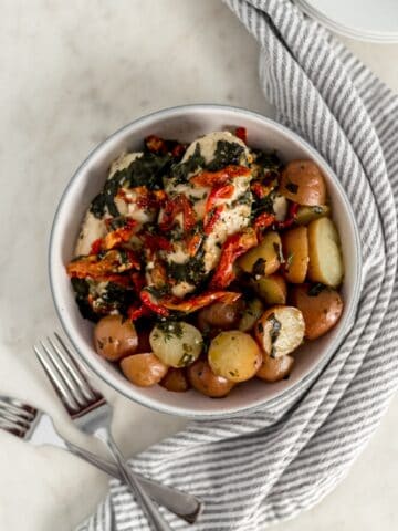 overhead view sun dried tomato chicken and potatoes in larger white bowl with two forks, plates, and napkin