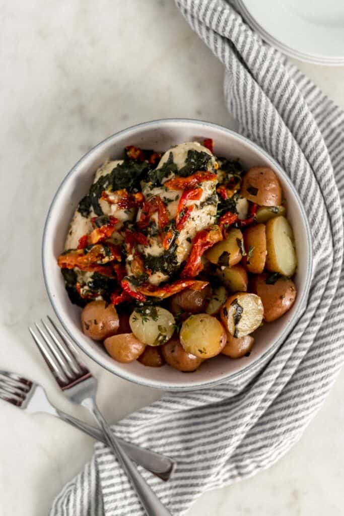 overhead view sun dried tomato chicken and potatoes in larger white bowl with two forks, plates, and napkin