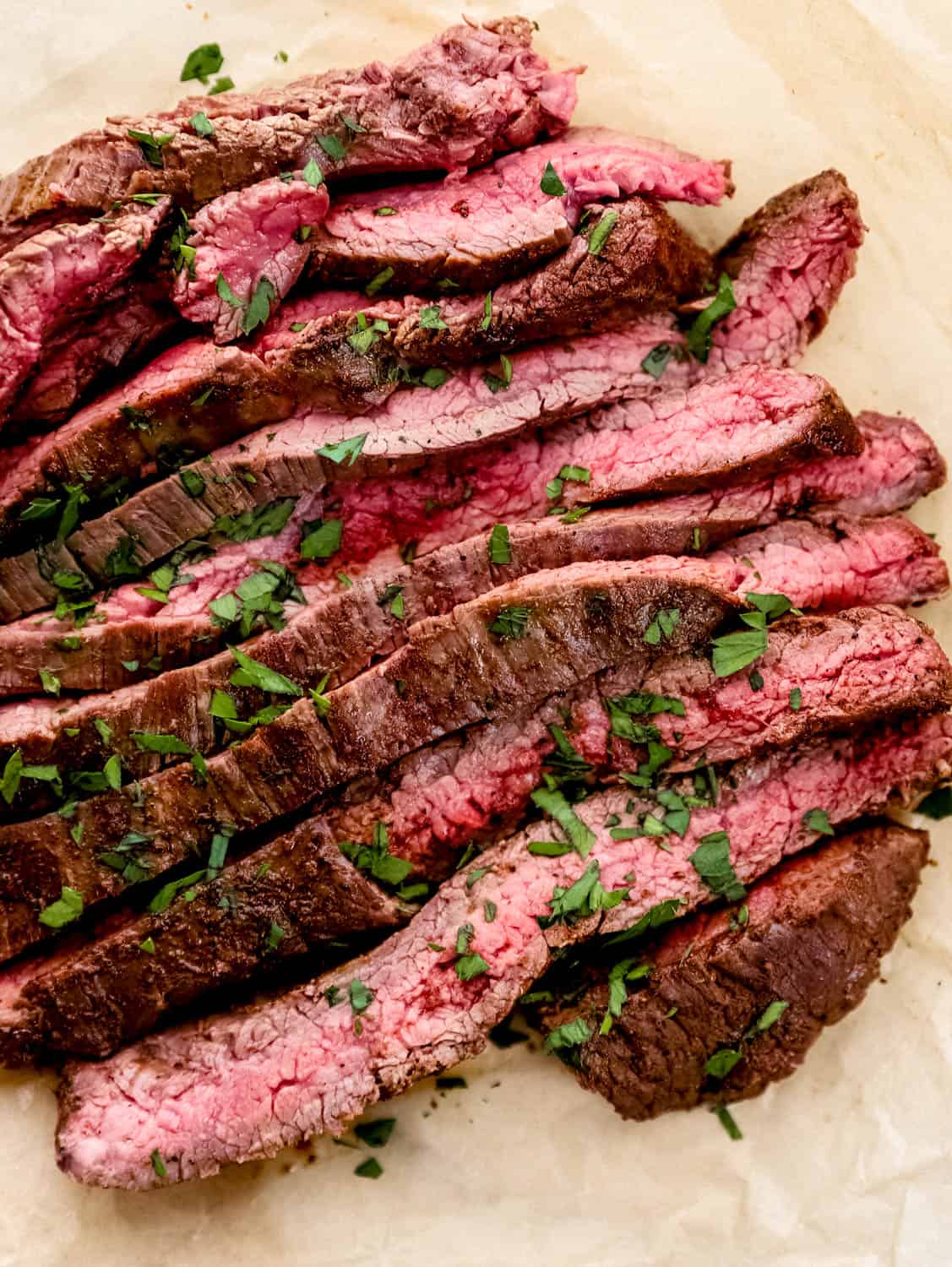Close up view of sliced steak topped with parsley on top of parchment paper.