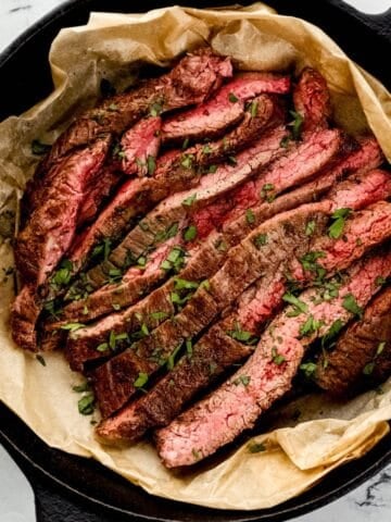 Overhead view of sliced steak topped with parsley in parchment lined cast iron skillet.