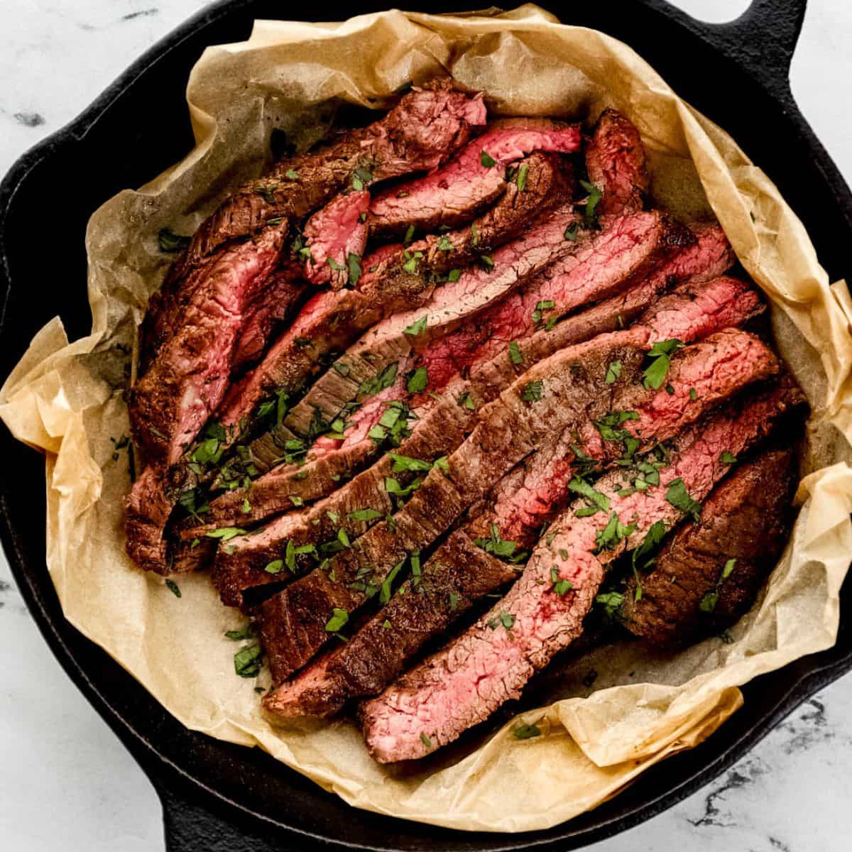 Overhead view of sliced steak topped with parsley in parchment lined cast iron skillet.