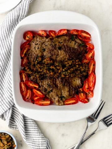 flank steak in square white baking dish with tomatoes, next to a cloth napkin, two forks, and white plates