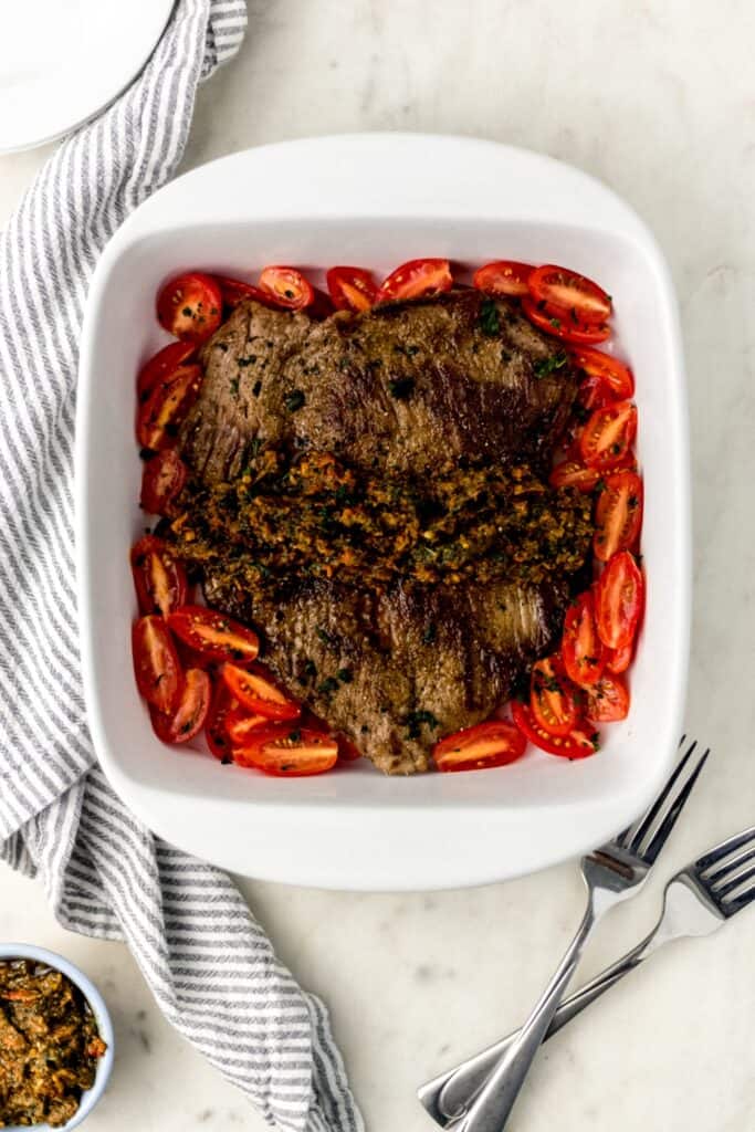 flank steak in square white baking dish with tomatoes, next to a cloth napkin, two forks, and white plates