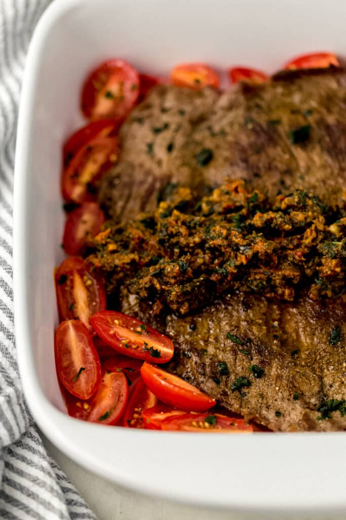 close up side view flank steak topped with pest in square white baking dish 