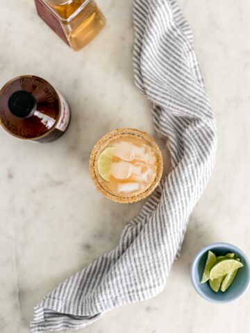 kombucha magarita in glass with lime wedge next to cloth napkin, small bowl with limes, and two bottles