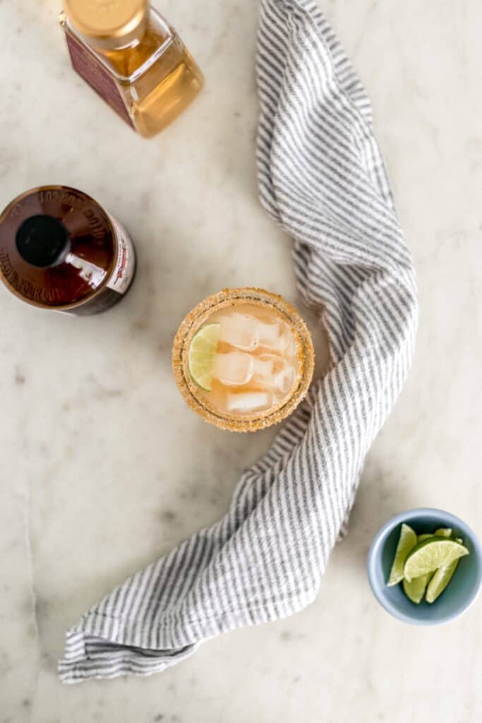 kombucha magarita in glass with lime wedge next to cloth napkin, small bowl with limes, and two bottles