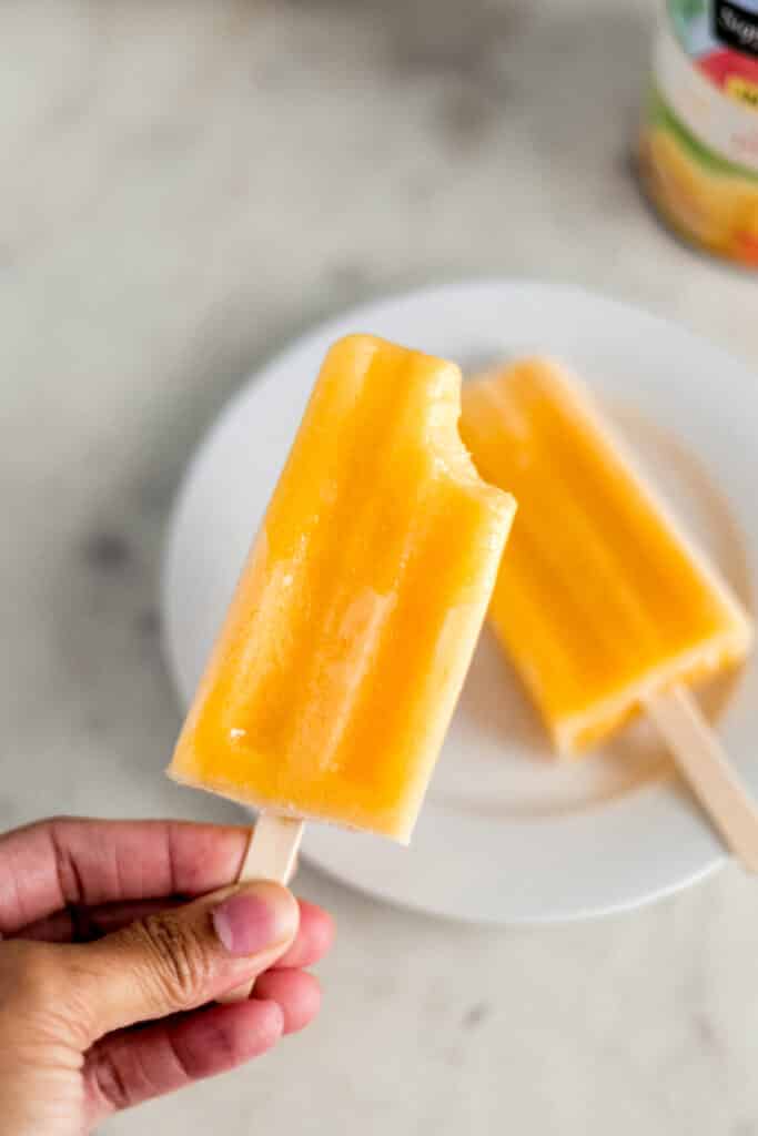 hand holding peach popsicle over white plate with another popsicle on it