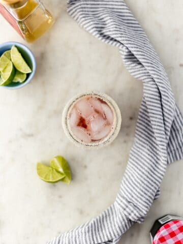 overhead view cherry margarita in sugar rimmed glass with limes, tequila, cherry preserves jar, and cloth napkin