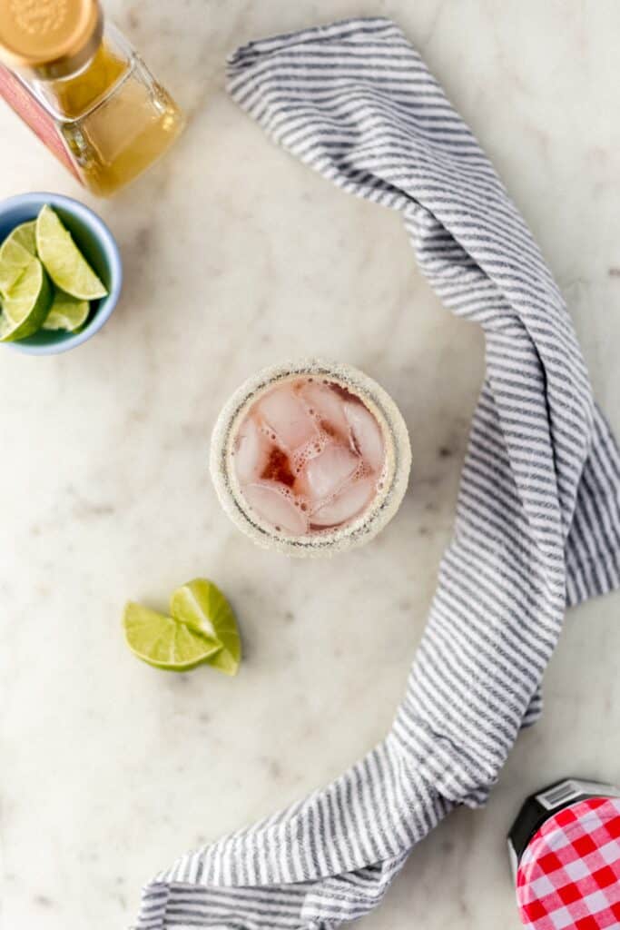 overhead view cherry margarita in sugar rimmed glass with limes, tequila, cherry preserves jar, and cloth napkin 