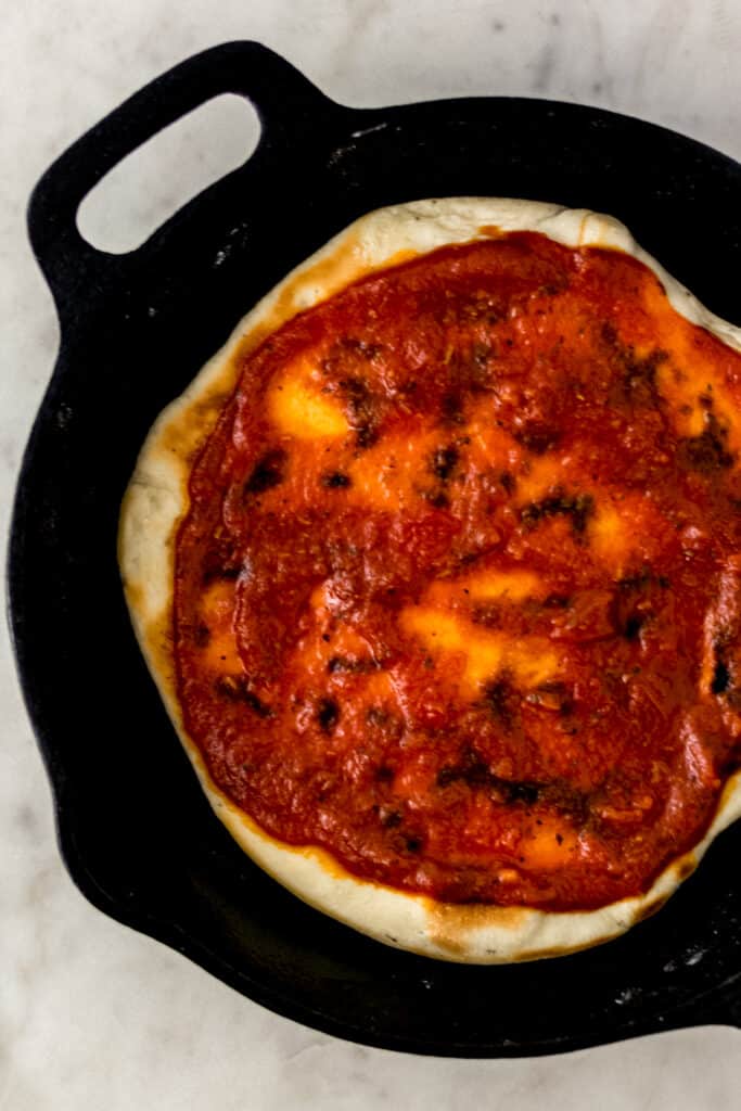 pizza crust topped with tomato sauce in cast iron skillet. 