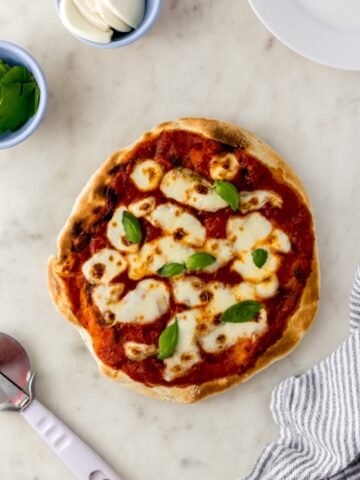 overhead view margherita pizza next to pizza cutter, small bowls with mozzarella and basil with cloth napkin.
