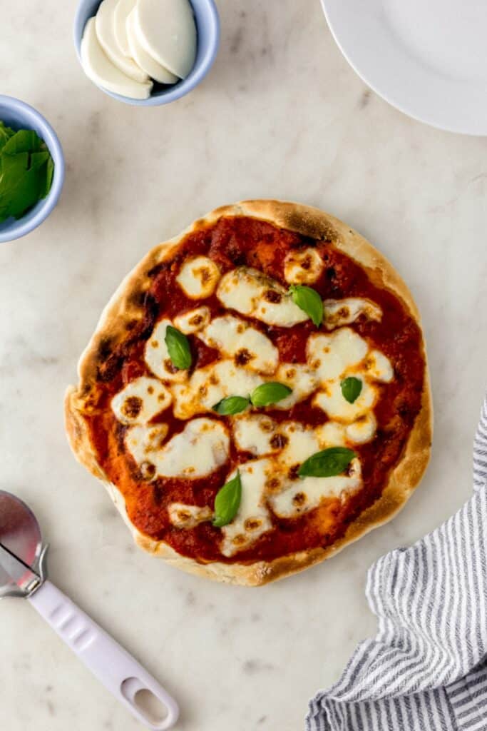 overhead view margherita pizza next to pizza cutter, small bowls with mozzarella and basil with cloth napkin. 