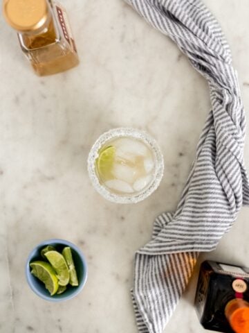overhead view of margarita in a glass rimmed with salt with bottles, limes, and cloth napkin