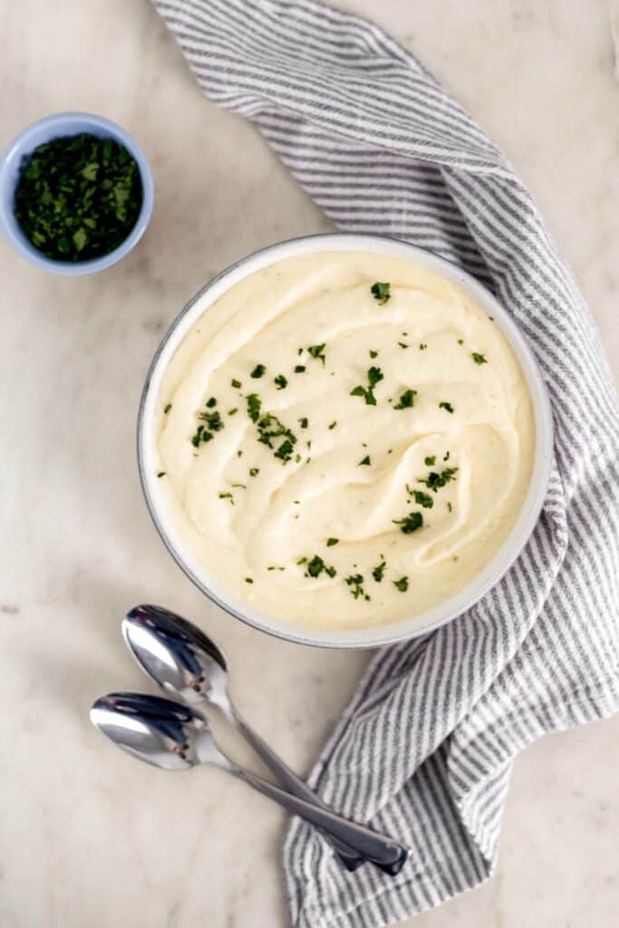 overhead view creamy mashed potatoes in bowl with spoons, cloth napkin, and small bowl with parsley. 