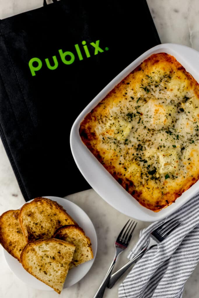 cheesy ravioli bake in square white baking dish with plate of garlic bread and grocery bag. 