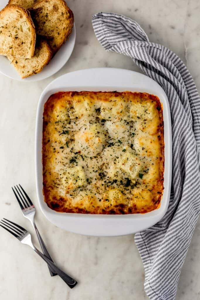 overhead of cheese ravioli bake in white baking dish with forks, cloth napkin, and white plate with garlic bread. 