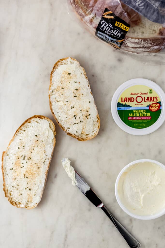 bread slices topped with butter next to a knife, butter container, and loaf of bread. 