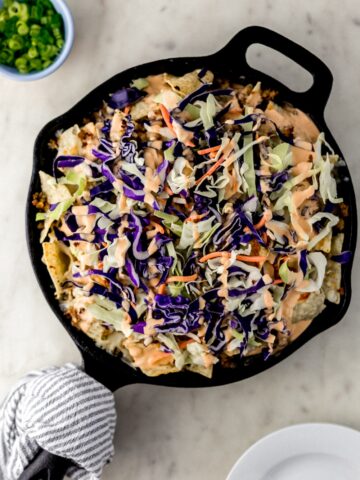 overhead view of pork nachos in cast iron skillet next to small white plates and small blue bowl with green onion