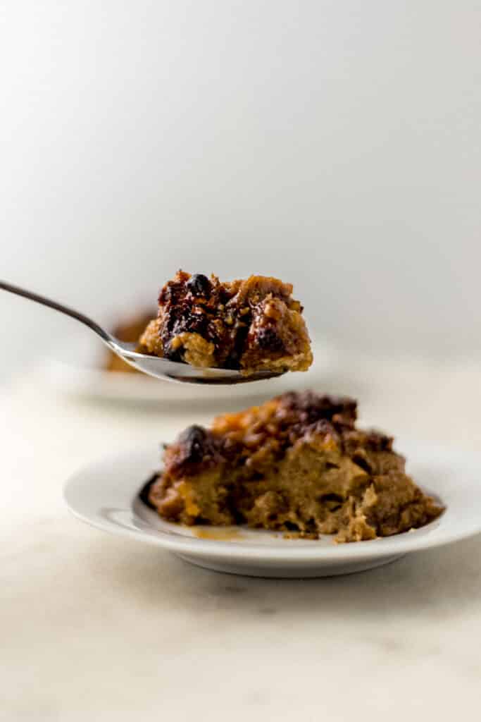 serving of peach bread pudding on white plate with hand holding spoon over it.