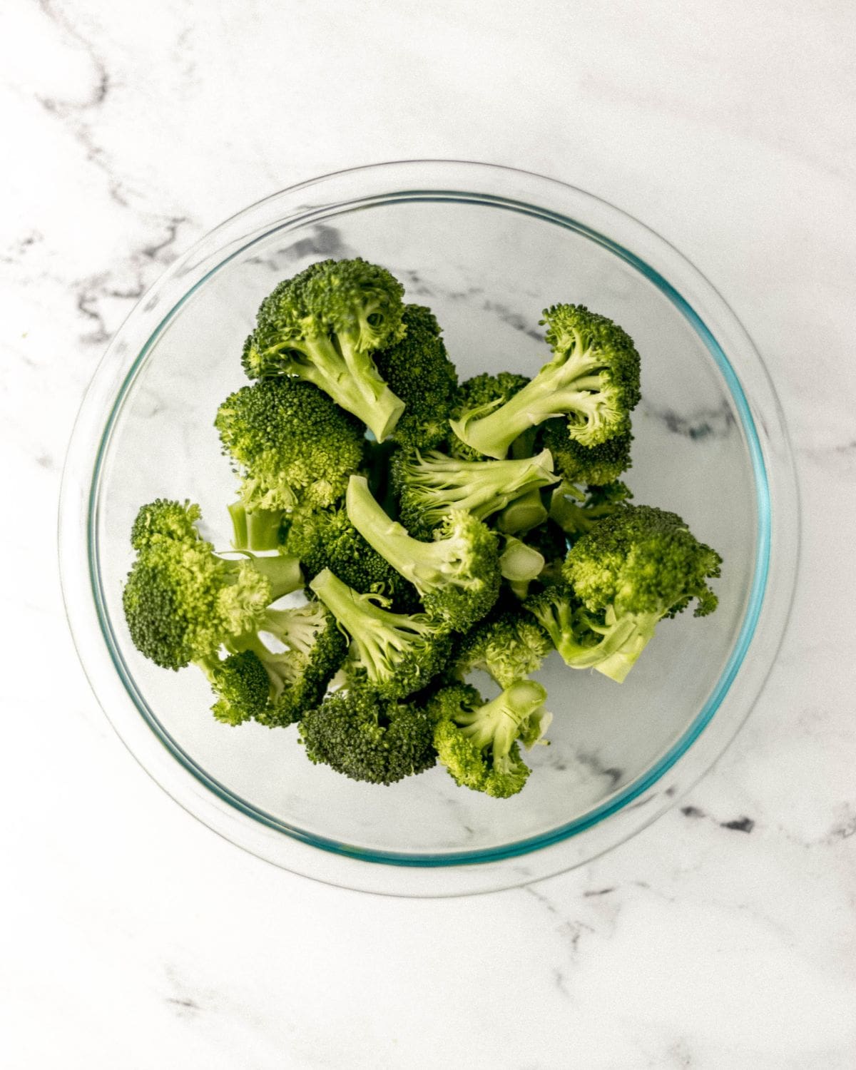 Fresh broccoli in large glass mixing bowl on marble surface. 