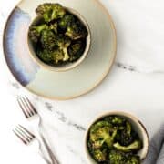 Overhead view of broccoli in two bowls by two forks on marble surface.
