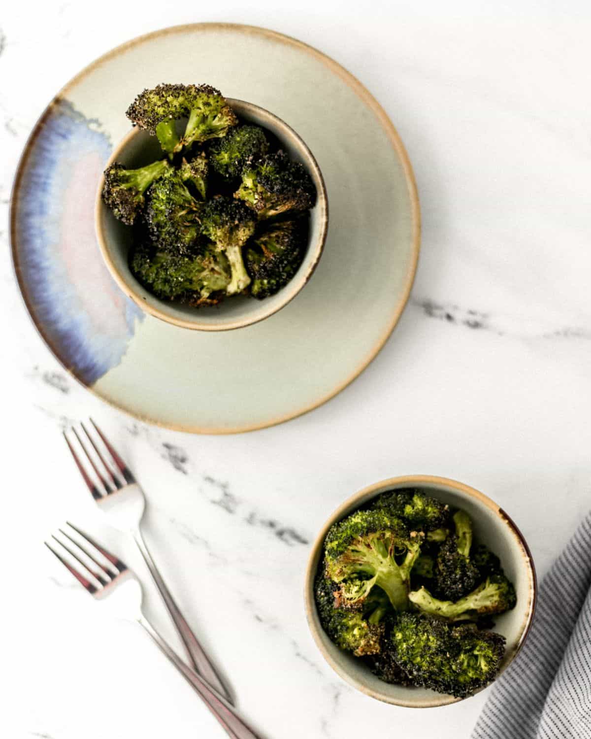 Overhead view of finished broccoli in two bowls by two forks on marble surface. 