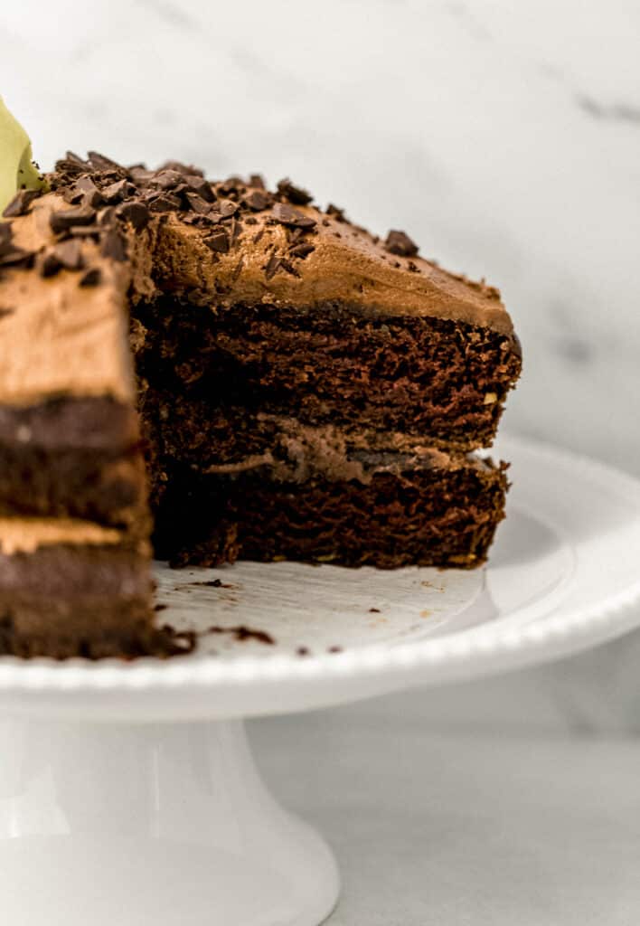 finished chocolate avocado cake with a slice cut out of it on white cake plate. 