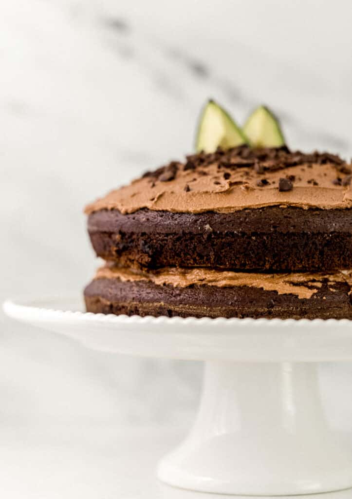 close up side view of frosted chocolate avocado cake on white cake plate. 