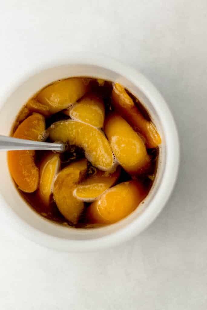 sliced peaches in bourbon glaze mixture in white bowl with spoon. 