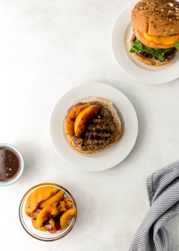 hamburger on white plate with small glass bowl of grilled peaches