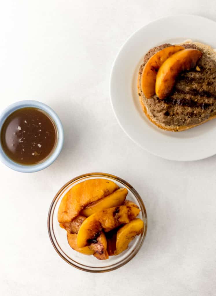 overhead view grilled peaches in glass bowl next to burger on white plate and sauce in a small bowl. 