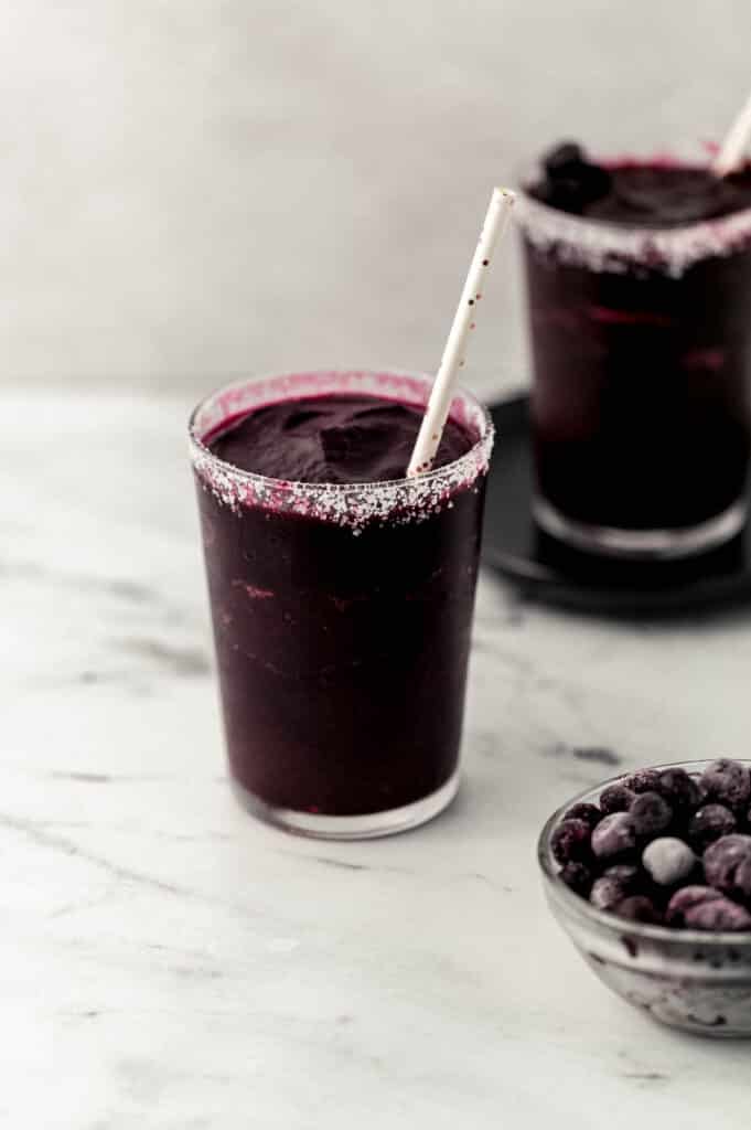 two salt rimmed glassed with blueberry margarita in them next to small glass bowl with frozen blueberries in it. 