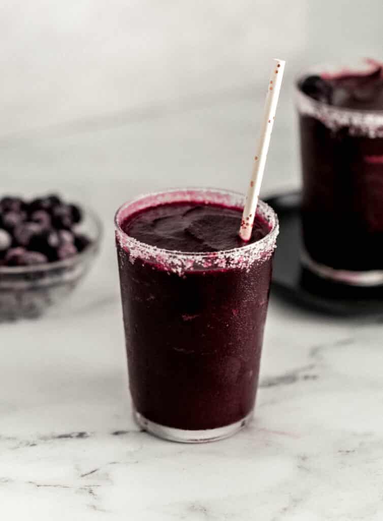 two frozen margarita in salt rimmed glasses, one on plate and next to glass bowl with frozen blueberries. 