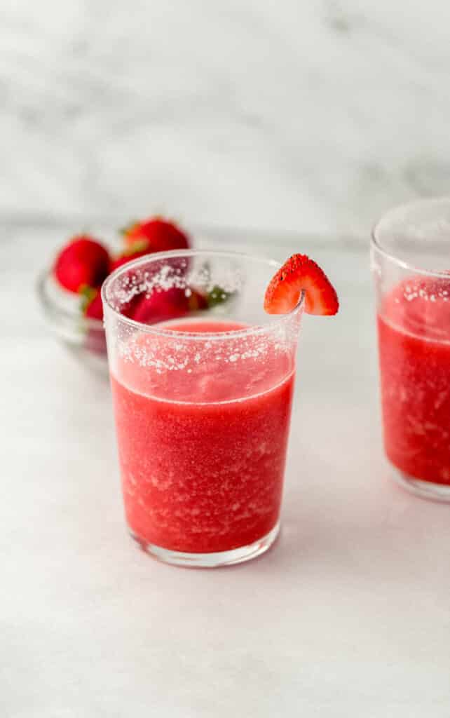 side view strawberry margarita in two glasses with small glass bowl of fresh strawberries in the background.