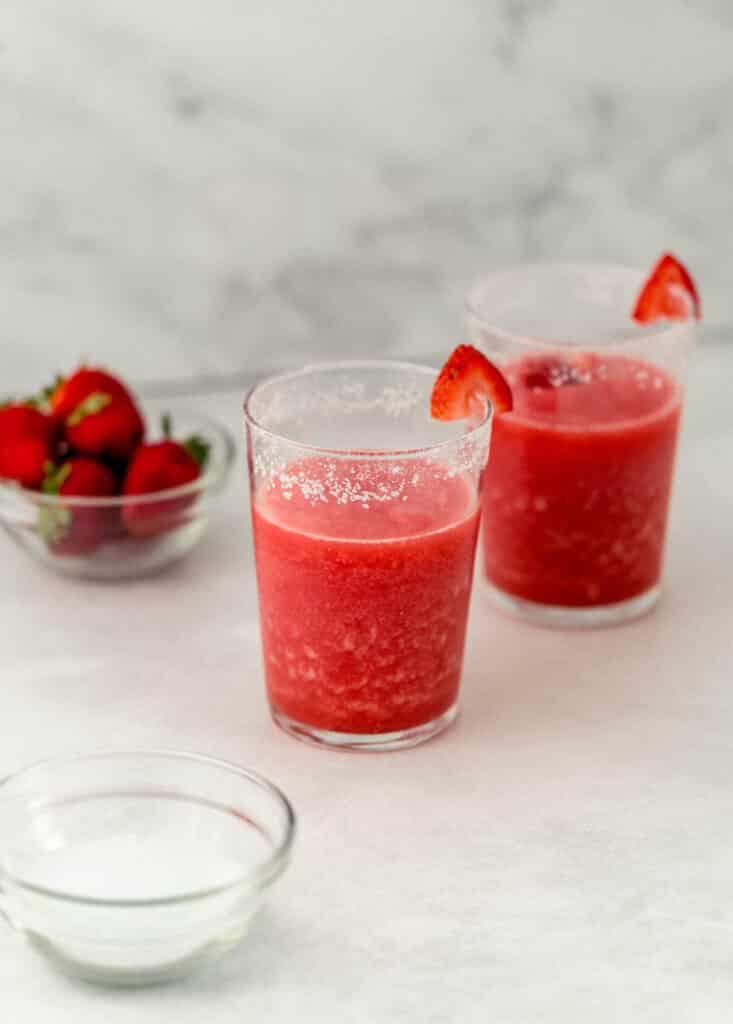 two glasses of frozen strawberry margarita with small bowls with salt and strawberries. 