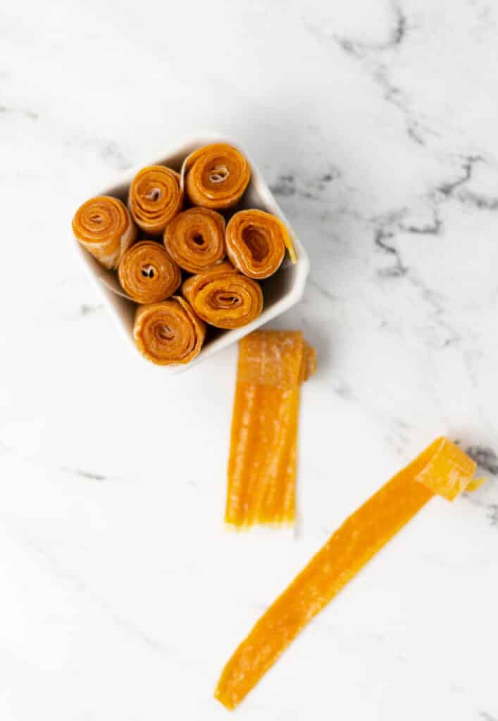 rolled up cling peach fruit leather in small square white bowl next to two unrollled pieces.