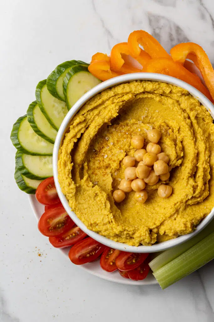 hummus in white bowl with fresh vegetables around it. 