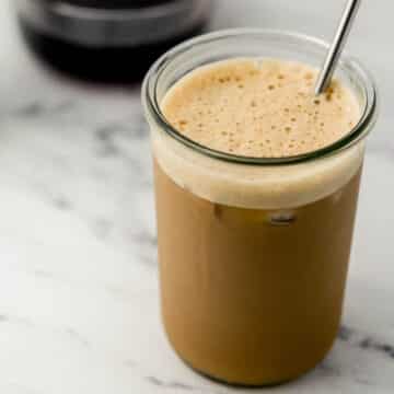 glass of iced shaken espresso with metal straw in it. Glass bowl of syrup in background.