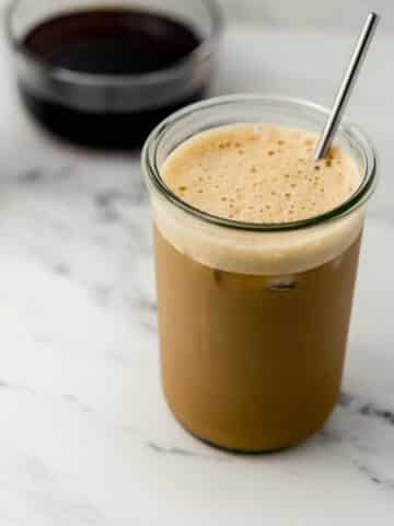 glass of iced shaken espresso with metal straw in it. Glass bowl of syrup in background.