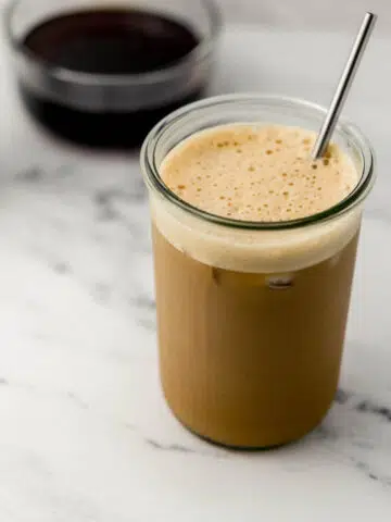 glass of iced shaken espresso with metal straw in it. Glass bowl of syrup in background.