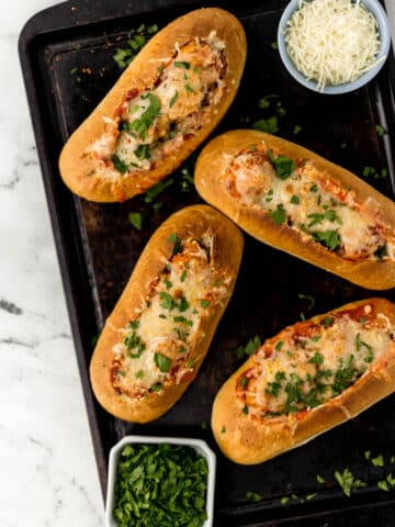 four meatball subs on a sheet pan topped with cheese and parsley