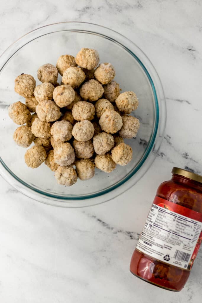 glass bowl with frozen meatballs in it next to jar of pasta sauce 