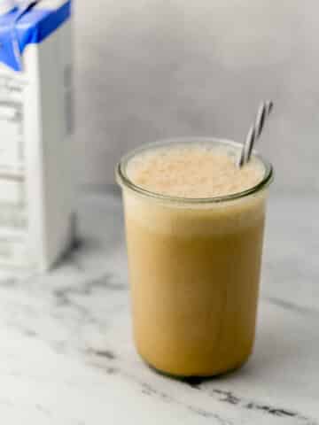 close up side view almond milk frappe in glass with straw with carton of almond milk in the background.