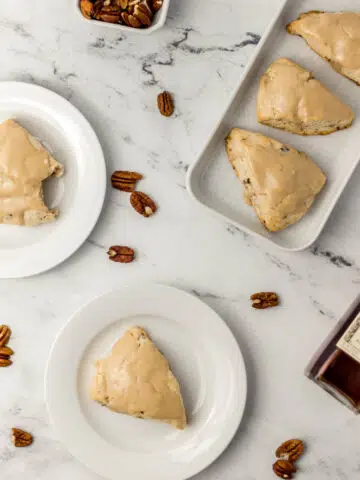 overhead view of maple pecan scones on white plates and platter with small container of pecans and jar of maple syrup