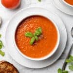 overhead bowl of tomato soup next to spoon, grilled cheese, fresh basil and tomatoes