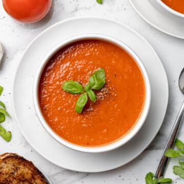 overhead bowl of tomato soup next to spoon, grilled cheese, fresh basil and tomatoes