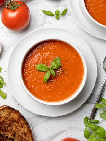 overhead bowl of tomato soup next to spoon, grilled cheese, fresh basil and tomatoes