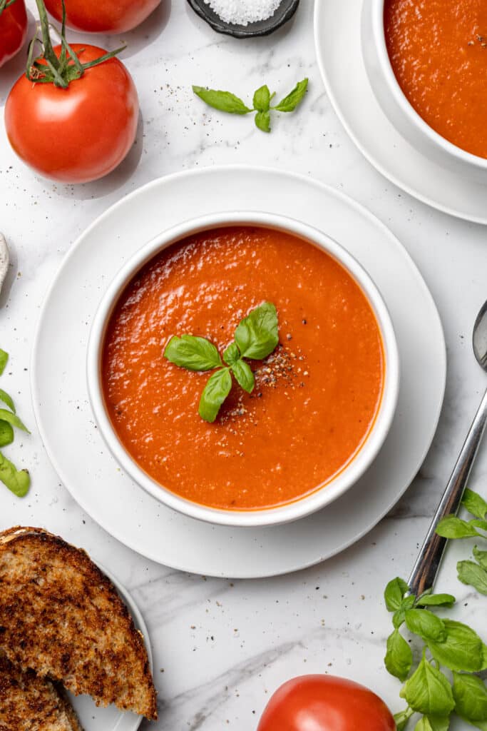 overhead view bowl of tomato soup topped with a piece of fresh basil next to a spoon, grilled cheese, fresh basil, and tomatoes.