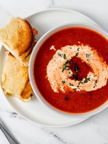 Overhead view of a bowl of tomato soup on top of a white plate with a sandwich on it beside a spoon.